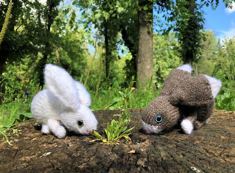 Two knitted bunny toys, one white and one brown, are positioned on a textured log amidst green foliage. The white bunny has pointed ears and a small pink nose, while the brown bunny is slightly hunched, both looking at a small patch of grass. The background features blurred green trees under a bright blue sky, creating a natural outdoor setting.