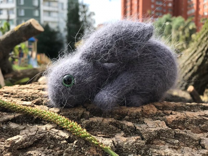 A fluffy, gray knitted bunny toy is positioned on a log, with its ears slightly perked and eyes made of green beads. The background features blurred greenery and buildings, suggesting an outdoor setting. The bunny's texture is soft and fuzzy, contrasting with the rough surface of the log.