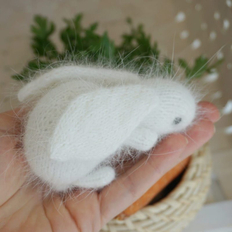 A hand holds a small, white knitted bunny toy with soft, fluffy texture, featuring long floppy ears and a rounded body. The bunny's eyes are represented by small stitched black dots, and it sits in a relaxed position. In the background, a woven basket filled with orange carrots is partially visible, along with greenery suggesting an indoor plant setting. The overall color palette is light and airy, with a focus on soft whites and natural earthy tones.