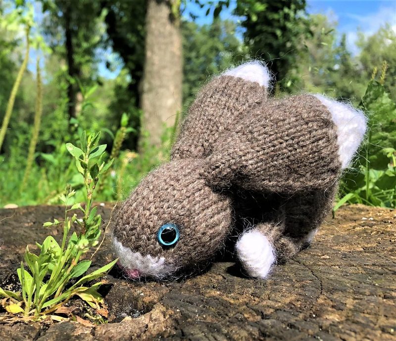 A knitted bunny toy is positioned on a wooden surface, with its body primarily in a brown yarn, accented by white fur details on the face and the underside. The bunny's blue button eye and small pink nose are visible. It appears to be sniffing a small green plant, surrounded by grass and trees in the background, creating a natural setting. The photo is brightly lit, emphasizing the bunny's texture and color contrast against the earthy tones of the wood and the vibrant greens of the surroundings.