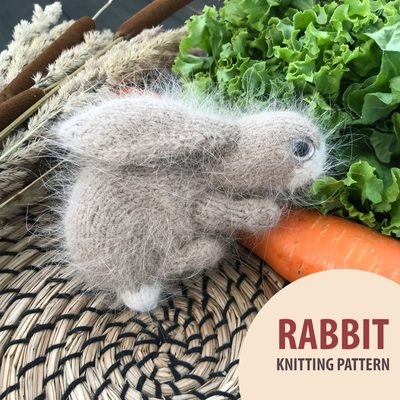 A small, fluffy bunny toy is positioned prominently in the foreground, showcasing light brown fur with a slightly fuzzy texture. The bunny has large, round eyes and long ears, appearing to be mid-hop. In the background, fresh green lettuce leaves and a bright orange carrot provide a colorful contrast, while a woven basket lies beneath. In the bottom right corner, there is a circular graphic that reads "RABBIT KNITTING PATTERN" in bold text. The overall scene has a natural, rustic aesthetic with