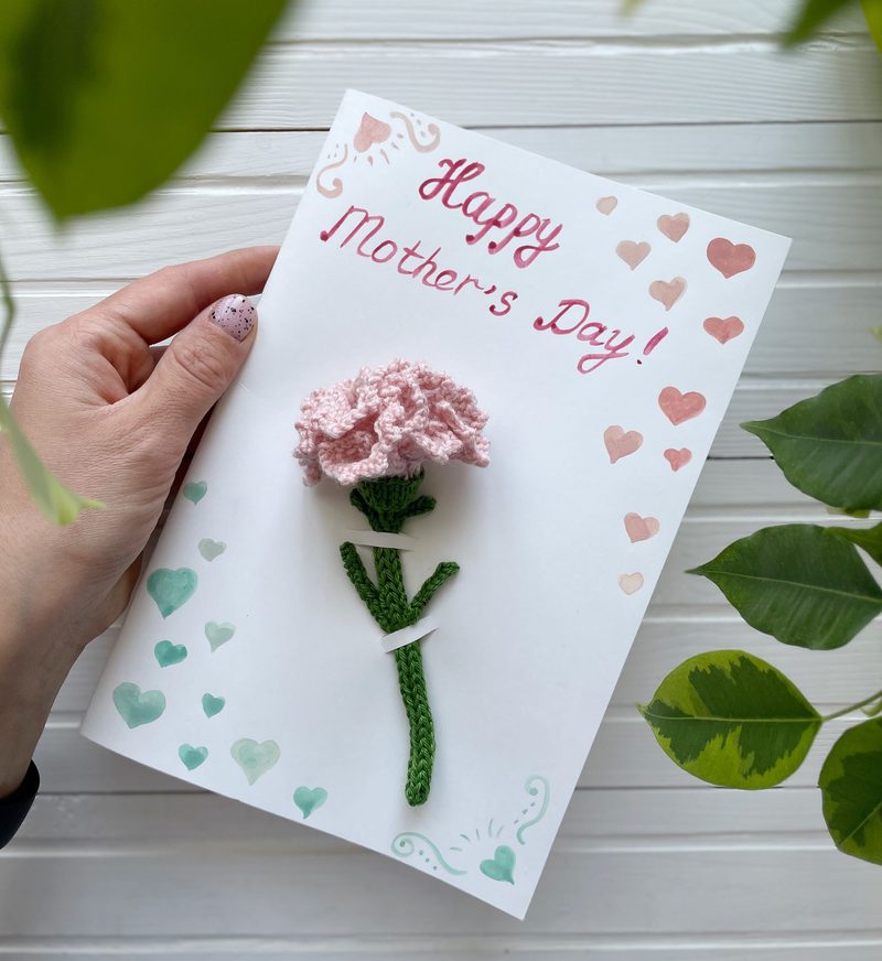 A person's hand holds a greeting card featuring a knitted pink carnation flower attached to a green stem. The card has red and mint green heart patterns around the edges and the text "Happy Mother's Day!" written in a whimsical font. The background includes green leaves, adding a touch of nature to the presentation.