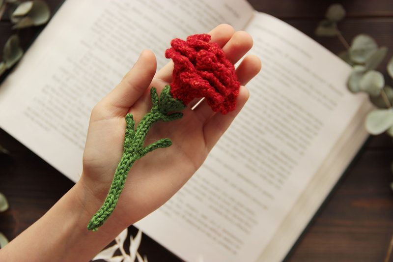 A hand holds a crocheted red carnation with a green stem against an open book with printed text, framed by soft, muted colors of a wooden surface and surrounding greenery.