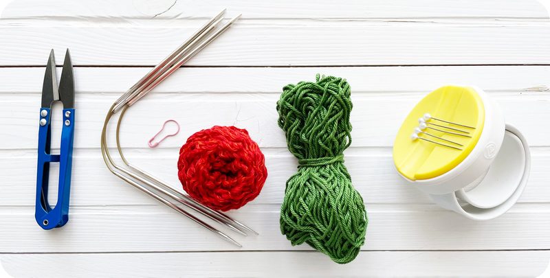 The image shows a flat lay arrangement of knitting supplies on a white wooden surface. On the left, there are blue scissors, followed by a red yarn ball, a green skein of yarn, and a set of metal knitting needles. A pink stitch marker is positioned next to the red yarn. On the right, there is a white yarn bowl with a yellow top and several white pins placed inside it. The overall composition is clean and organized, showcasing the vibrant colors of the yarn against the neutral background.