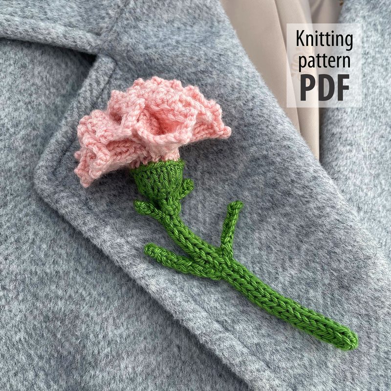 The image features a knitted carnation flower in soft pink at the top of a green stem with leaves. The flower is textured, resembling the layered petals of a real carnation. The background shows a gray textured fabric, likely a coat. To the right, there is a text overlay that reads "Knitting pattern PDF." The overall composition highlights the detailed craftsmanship of the flower against the neutral fabric backdrop.