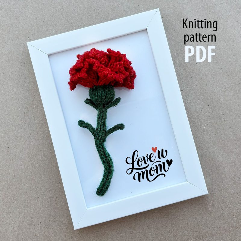 A framed image of a knitted red carnation flower with a green stem and leaves, against a neutral beige background. The flower is prominently displayed at the top, while the base features the text "Love u mom" in a cursive font, accompanied by a small heart, and a label on the side indicating "Knitting Pattern PDF." The frame is white, providing a clean contrast to the colorful knitted design.
