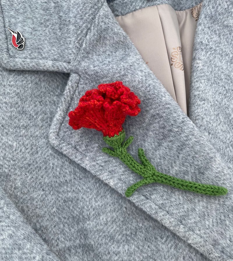 A close-up image of a gray coat featuring a crocheted red carnation flower pin, attached at the lapel. The flower has textured petals and a green stem, contrasting sharply with the coat's fabric. The background is blurred, focusing attention on the flower pin's intricate details and vibrant colors.