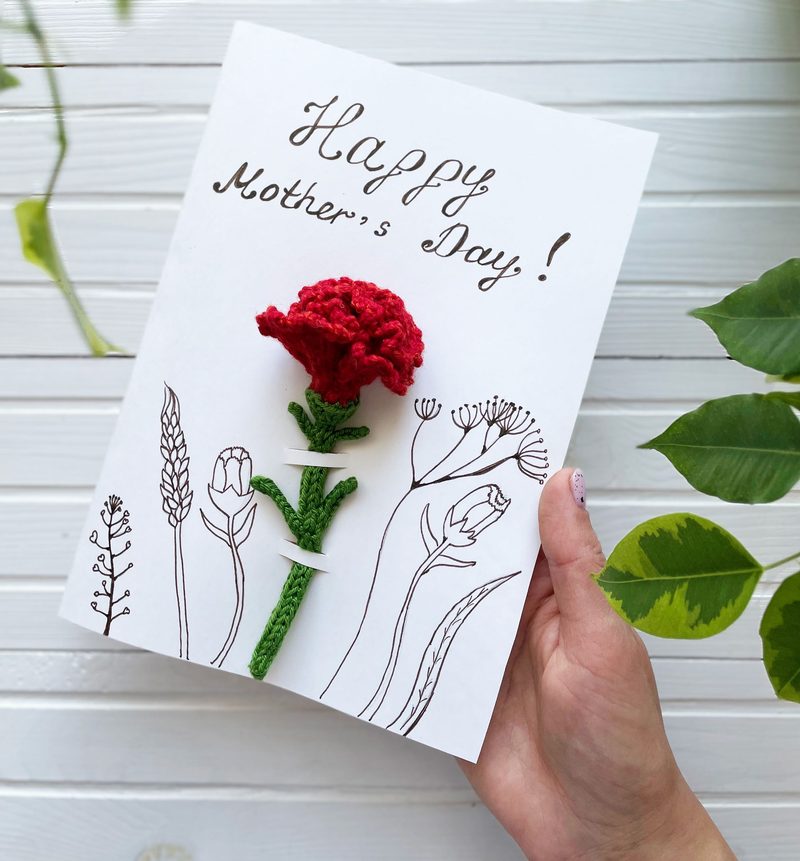A hand holding a card featuring a red crocheted carnation flower attached to a white background, with simple line drawings of various flowers and plants. The card includes the text "Happy Mother's Day!" written in a decorative script at the top. The background consists of white wooden planks, with green leaves partially visible at the edges, adding a natural touch to the composition.