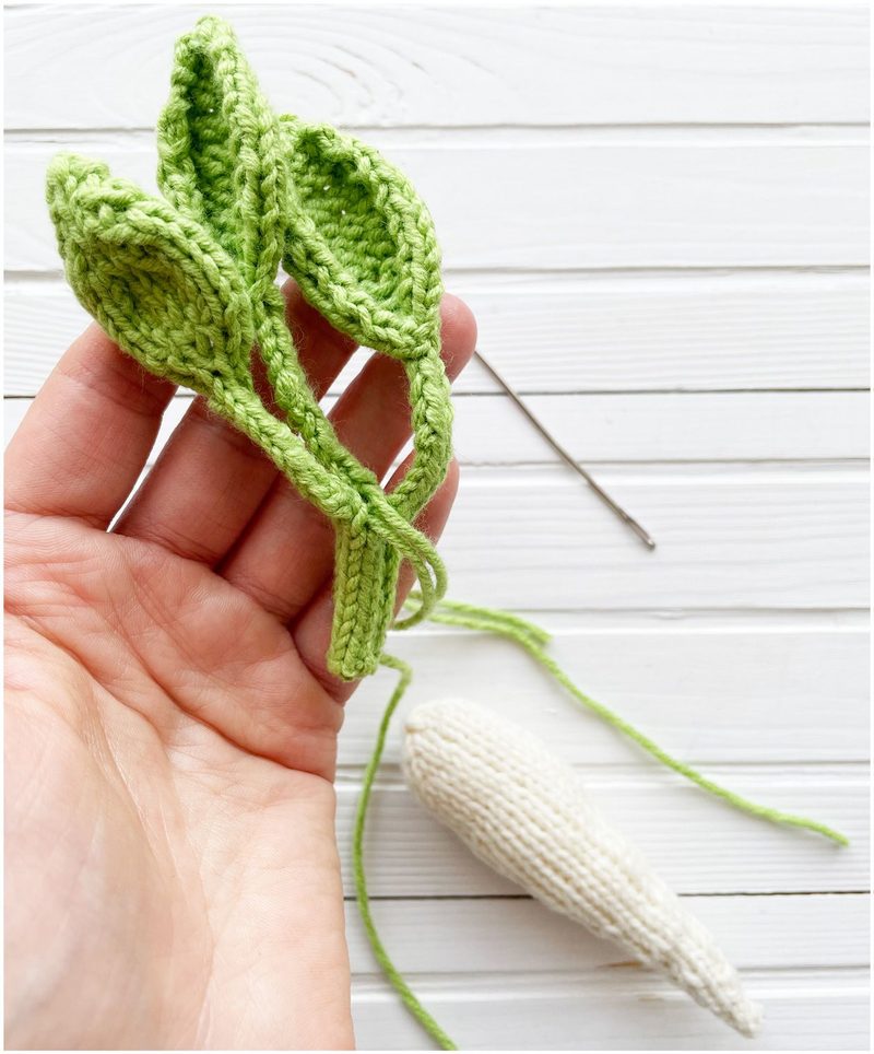 A hand holds three leaf shapes made of bright green yarn, displaying a textured knitting pattern, while a white, elongated carrot shape is partially visible below, resting on a light wooden surface. A silver knitting needle is positioned nearby, and the background features a white wood texture.
