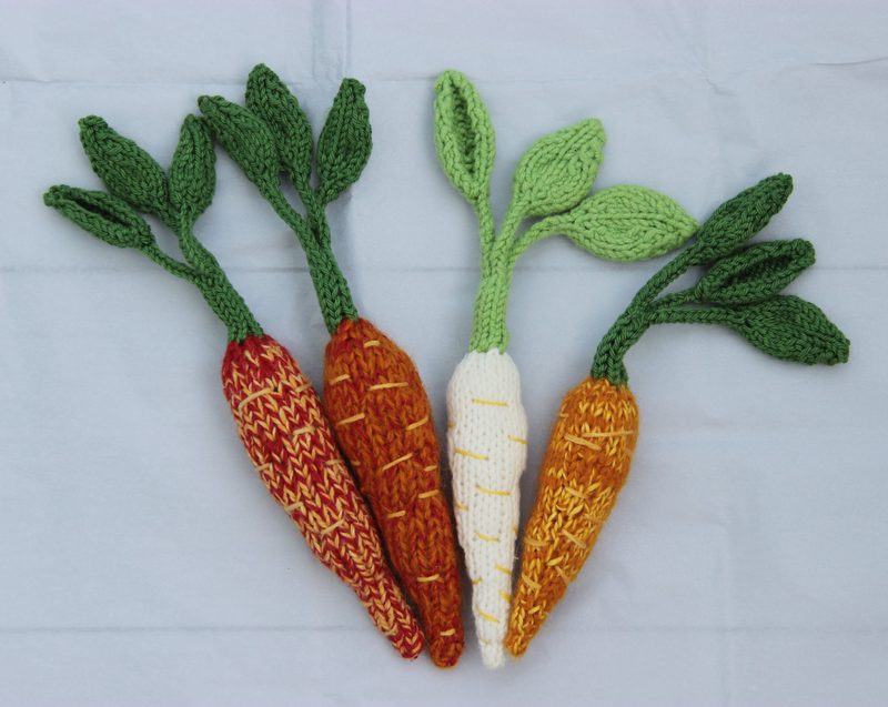 Image features four knitted amigurumi carrots in various colors and patterns. The leftmost carrot has an orange and yellow zigzag design, next to it is a solid orange carrot, followed by a white carrot with yellow stitching, and finally, a carrot with a textured orange and white pattern. Each carrot has green knitted leaves extending from the top. The background is a light gray fabric that contrasts with the vibrant colors of the carrots.
