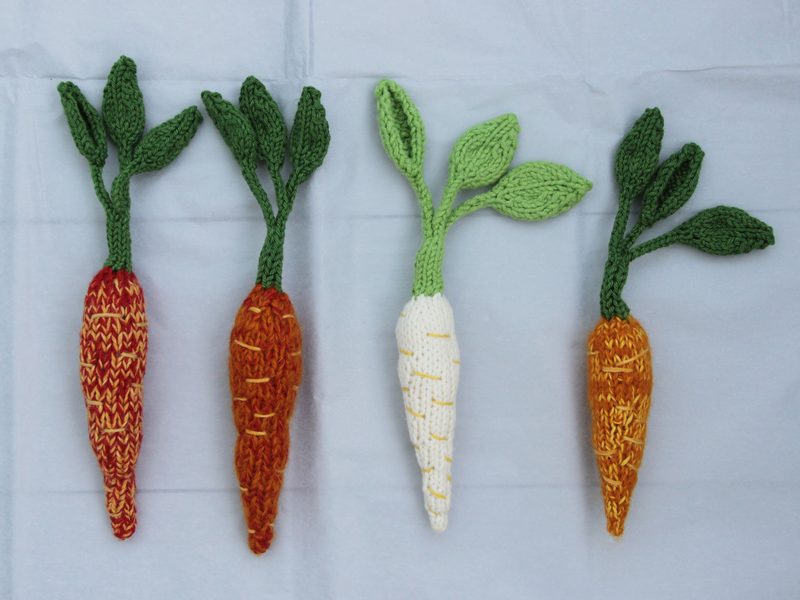Four knitted amigurumi carrots are displayed on a light-colored background. The carrots vary in color and design: two are orange with textured patterns, one is white with light yellow accents, and one is a lighter orange. Each carrot has vibrant green leaves that are crafted in different shades, arranged to give a lively appearance. The patterns are intricate, showcasing a blend of yarn textures and colors. The overall composition is balanced, with the carrots lined up horizontally.