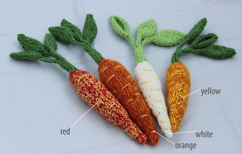 The image shows four crocheted amigurumi carrots arranged side by side. Each carrot has a different color: red, orange, white, and yellow, with textured patterns on the red and orange ones. Each carrot features green leaf shoots at the top, crafted with yarn that has a vibrant green hue. The background is a light, neutral surface that complements the colorful carrots. The overall composition is flat lay, highlighting the details of the yarn and the distinct colors of each carrot.
