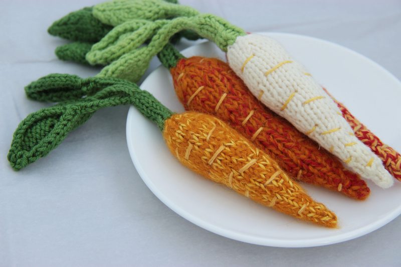A white plate displays three amigurumi carrots: one white, one orange, and one darker orange, all with textured yarn detailing. Each carrot features vibrant green leaves made of knitted fabric. The background is a light surface, providing a neutral contrast to the colorful carrots.