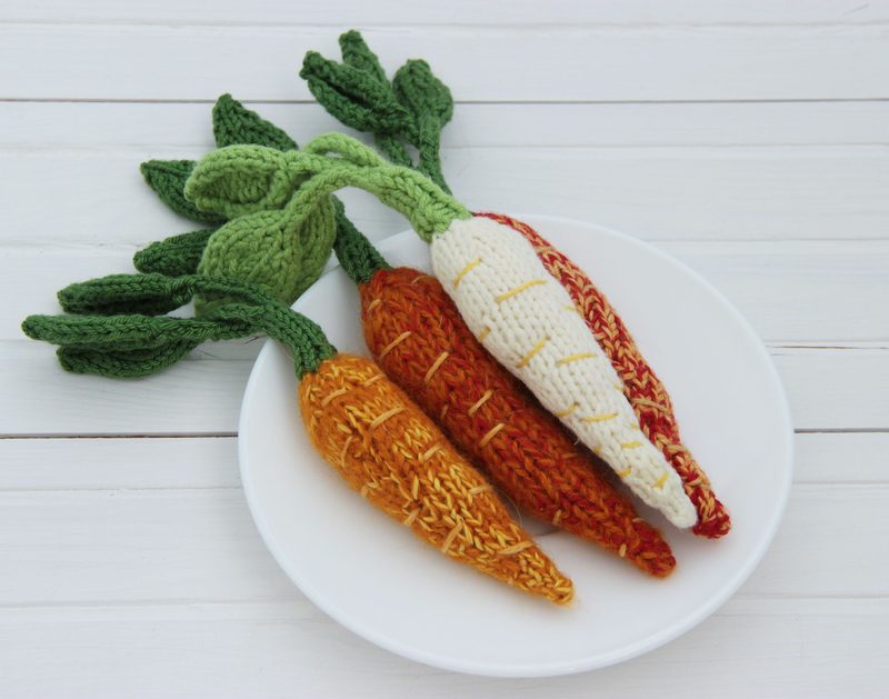 A white plate displays four crocheted amigurumi carrots, each with a distinct color and design; two are orange, one is yellow with orange accents, and one is white with an orange stripe. The green tops are detailed with textured leaves, adding a realistic touch. The background is a simple wooden surface, enhancing the vibrant colors of the carrots.