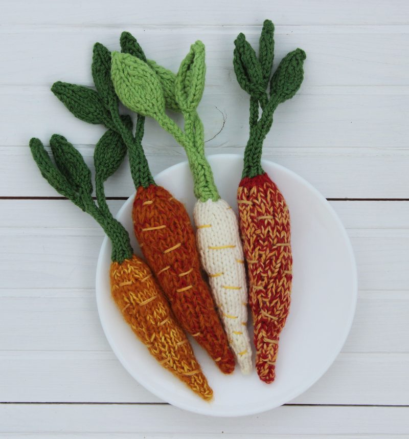 A white plate displays four knitted amigurumi carrots in various colors, including orange, cream, and a reddish-brown. Each carrot features green leafy tops, with the textures and stitches of the knitting clearly visible. The background is a light wooden surface, enhancing the vibrant colors of the carrots.