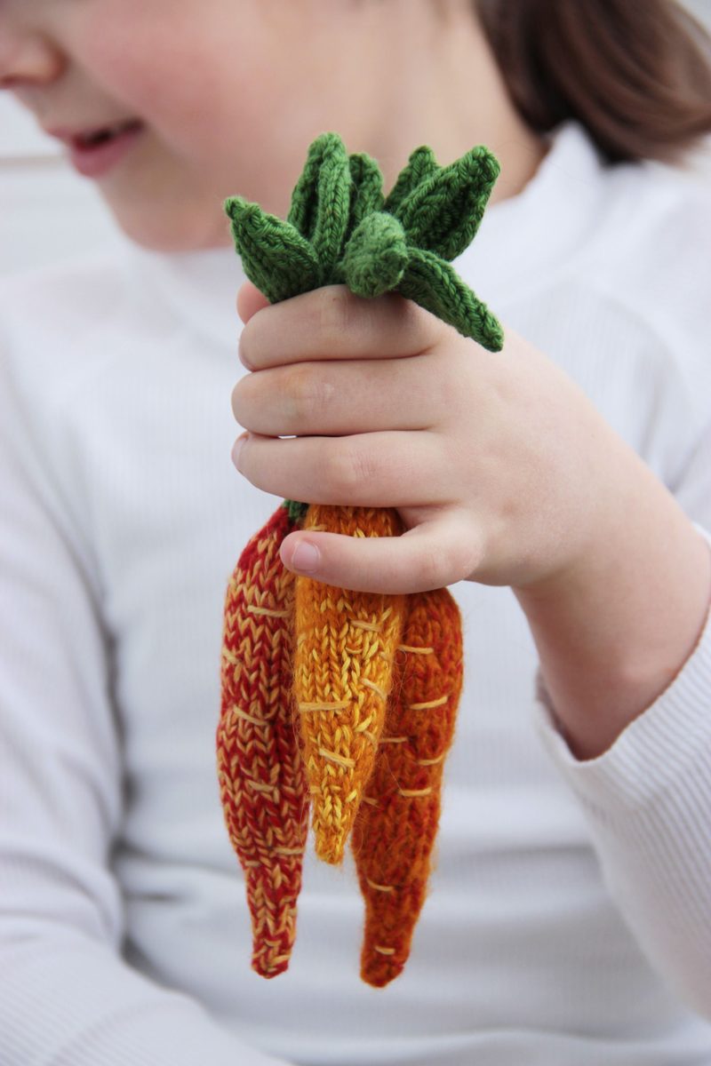 A child’s hand holds three knitted amigurumi carrots in varying shades of orange and yellow, with green leaves at the top. The carrots have textured detailing and are arranged in a bunch. The child is wearing a white long-sleeve shirt, but their face is not visible, and the background is softly blurred.