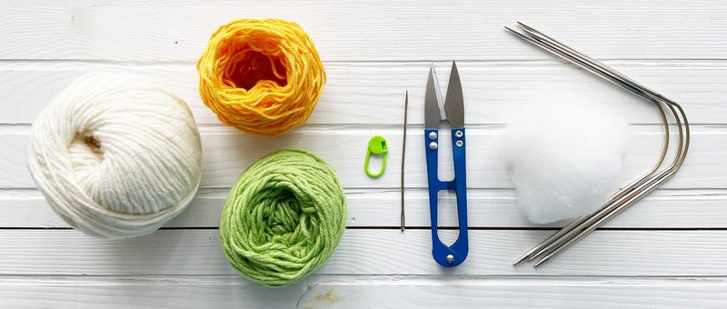 A flat lay of crafting materials for an amigurumi project, including three skeins of yarn in white, orange, and green, a pair of silver scissors, a blue crochet hook, a green stitch marker, a sewing needle, and a small bundle of white stuffing, all arranged on a white wooden surface.