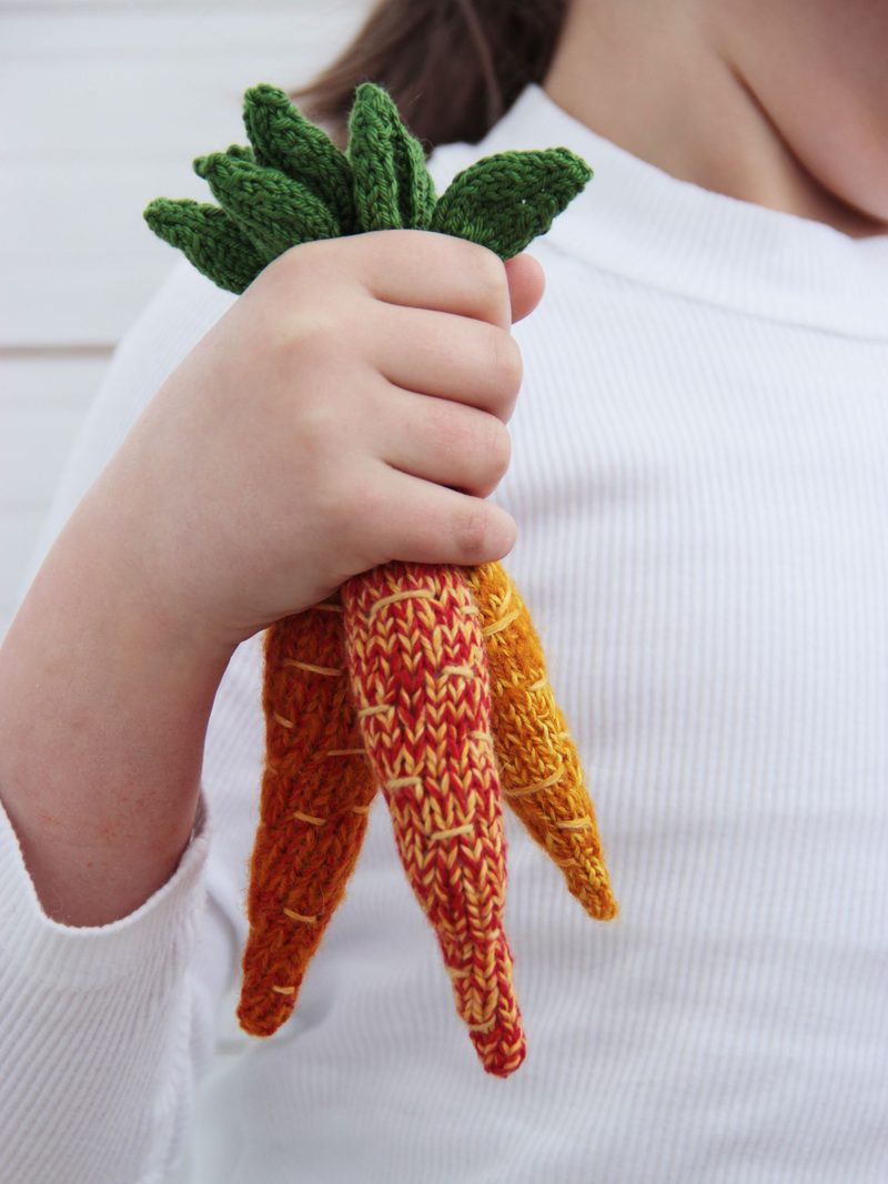 A child's hand holds a bunch of three knitted amigurumi carrots, featuring vibrant colors ranging from orange to yellow for the carrot bodies and deep green for the leaf tops. The carrots are intricately textured with visible stitching patterns, and the child is wearing a white long-sleeve shirt. The background is lightly blurred, focusing attention on the carrots.