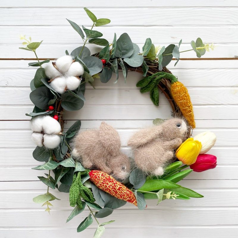 A circular wreath made of green foliage, cotton branches, and flowers, featuring two small, fluffy rabbits. One rabbit is positioned on the left, and the other on the right. The wreath includes colorful elements such as yellow tulips, a red amigurumi carrot, and an amigurumi corn, creating a vibrant, spring-themed display against a light wooden background.