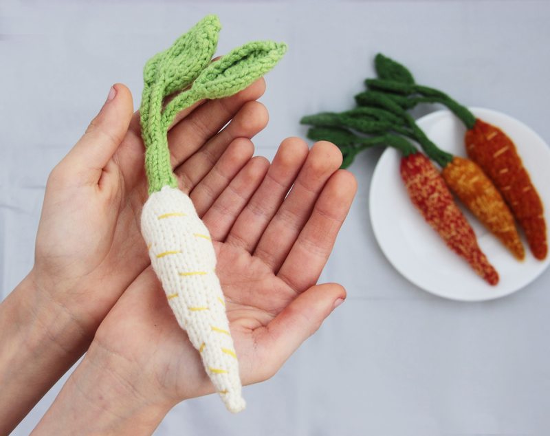 A person’s hands hold a white and green knitted carrot with yellow stitching, while in the background, three additional carrots in varying shades of orange and green are arranged on a white plate. The hands are positioned in the foreground against a neutral gray background, emphasizing the texture of the knitted carrot and the contrast with the colorful ones behind it.