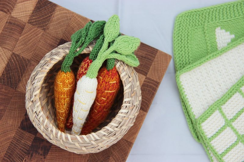 A woven basket containing three amigurumi carrots with distinct colors: one orange, one yellow, and one white, each topped with green, textured leaves. The basket is placed on a wooden cutting board, and a knitted green and white blanket, featuring a geometric pattern, is partially visible in the upper corner of the image. The overall scene has a warm and cozy aesthetic, with soft colors and textured materials.