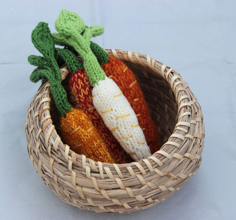 A woven basket filled with three knitted amigurumi carrots, featuring two orange carrots and one white carrot, all with green leafy tops. The carrots are detailed with texture, showcasing stitches in varying shades of orange and green, while the basket is positioned centrally on a light gray background.