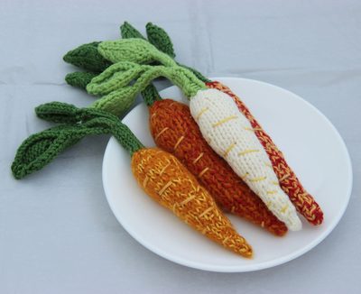 A white plate features four knitted amigurumi carrots in varying colors: two orange carrots, one with textured stitching, one with a smooth finish, a cream-colored carrot, and one with a mix of orange and red stripes. Green leaves protrude from the tops of the carrots, showcasing a blend of light and dark greens. The background is a soft gray, providing contrast to the vibrant colors of the carrots.