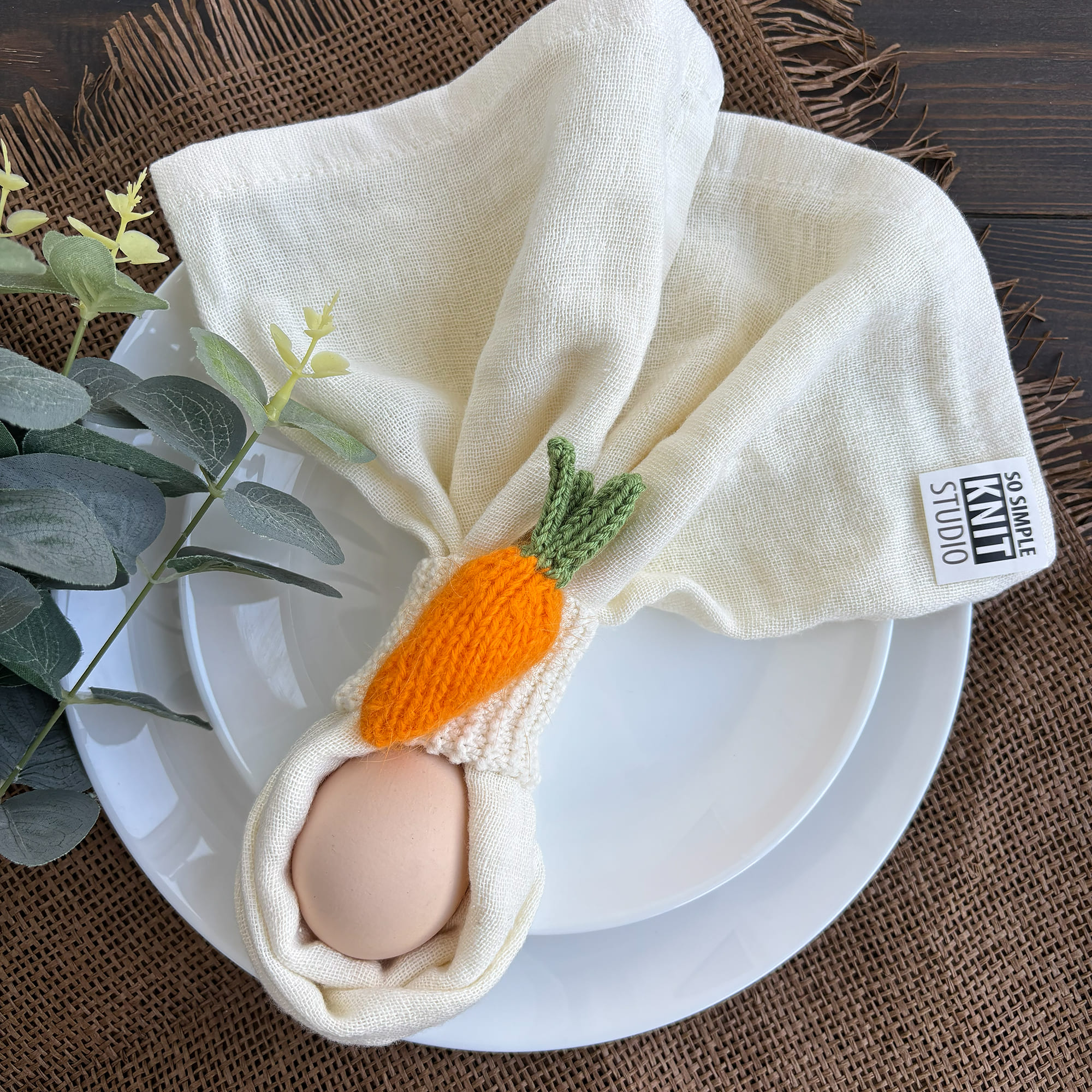 A white cloth napkin held by a knitted carrot napkin ring on a white plate; an egg is tucked inside the folded napkin, with decorative greenery on a textured fabric background nearby.