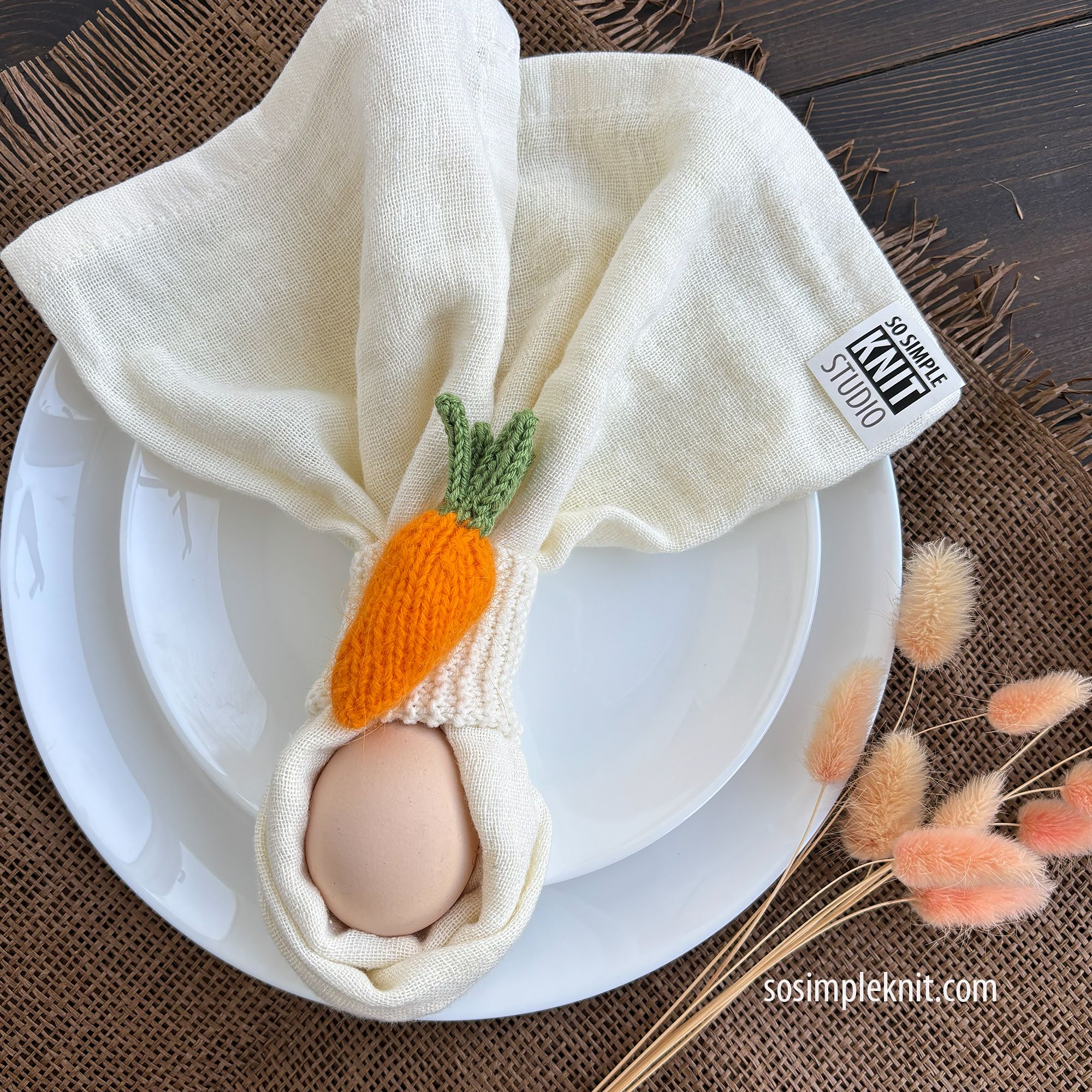 A white plate with a light cream-colored napkin, wrapped with a knitted carrot-shaped napkin ring. This is a beginner-friendly knitting pattern made with needles.