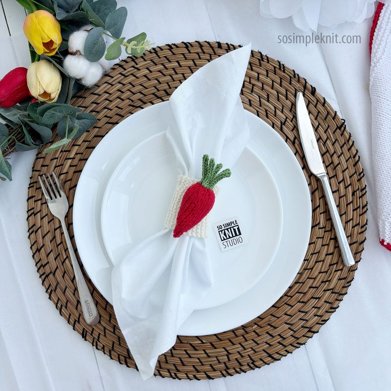 A white dinner plate is centered on a woven placemat. A knitted carrot napkin ring, featuring orange and green yarn, holds a white napkin neatly arranged on the plate. A fork and knife are placed beside the plate, and in the background, there are colorful artificial flowers and greenery. The image is bright and well-lit, emphasizing the texture of the napkin ring and the simplicity of the table setting.