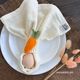 A white plate with a light cream-colored napkin, wrapped with a knitted carrot-shaped napkin ring. This is a beginner-friendly knitting pattern made with needles.