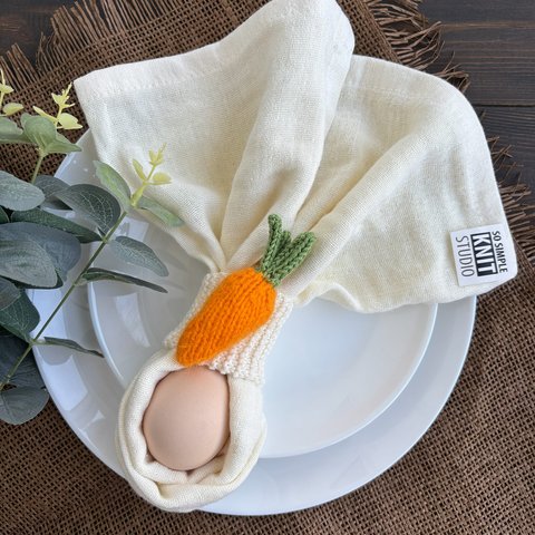 A white cloth napkin held by a knitted carrot napkin ring on a white plate; an egg is tucked inside the folded napkin, with decorative greenery on a textured fabric background nearby.