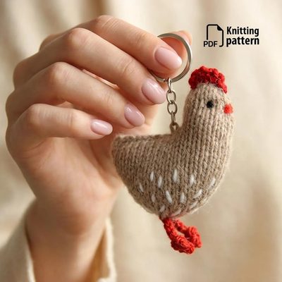 small, knitted chicken keychain made of beige yarn with white speckles, a red comb, and a red tail. The chicken is round and appears soft, suspended by a silver keyring. The background is softly blurred, focusing on the chicken and the hand holding it. The text "PDF Knitting pattern" is visible in the top right corner in simple, modern font.