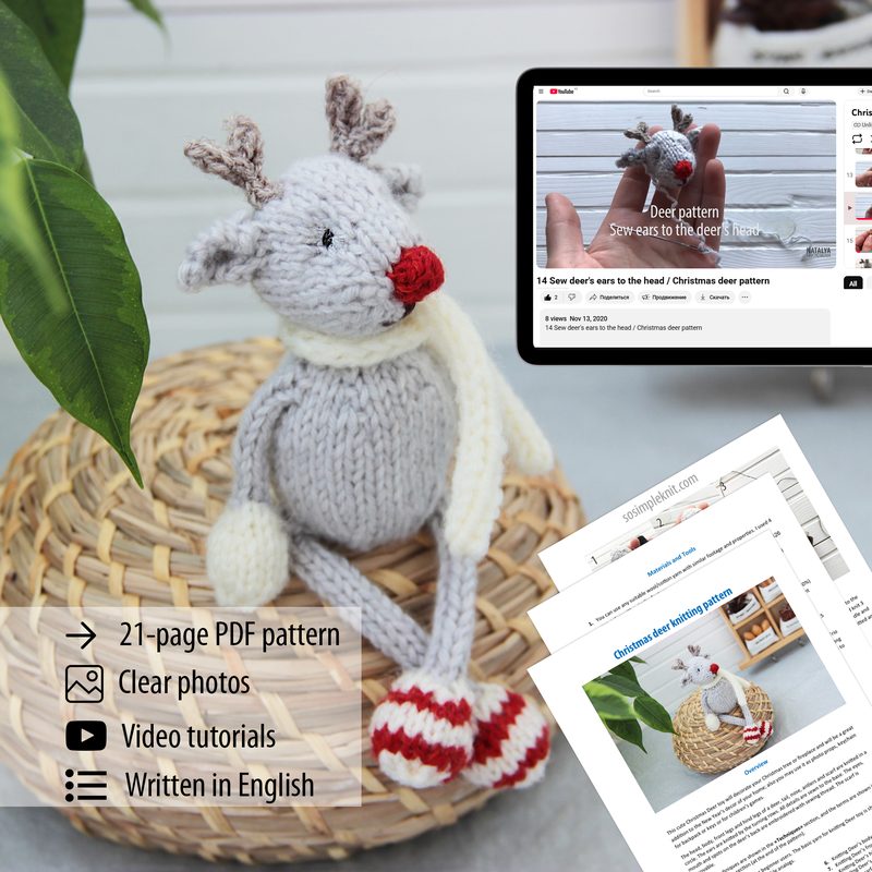 A knitted reindeer toy is seated on a woven basket, featuring a gray body, white antlers, a red scarf, and striped red and white booties. In the background, an electronic tablet displays a webpage with the title "Deer pattern" and images of the knitting pattern. Next to the tablet, printed pages of the pattern are visible, along with icons indicating features of the pattern, such as a PDF format and video tutorials. The setting includes a green plant partially visible in the foreground.