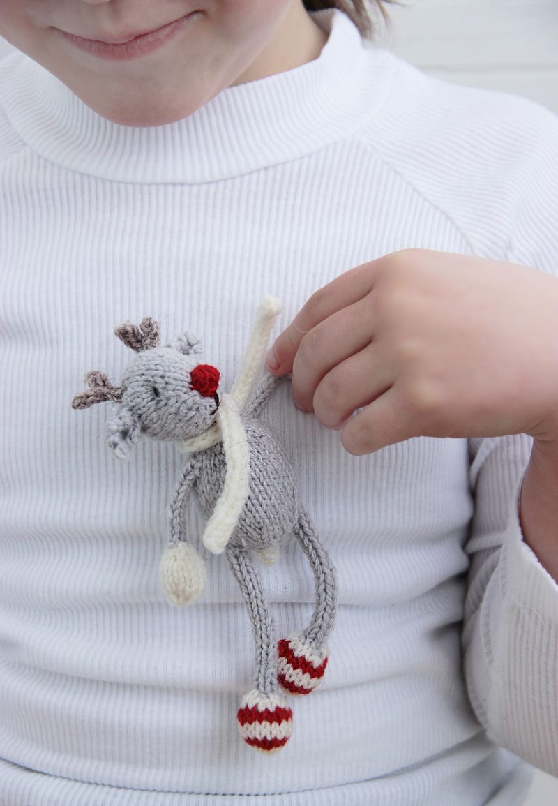 A child in a white long-sleeve shirt holds a small knitted reindeer toy. The reindeer, in gray with a red nose, features small antlers and wears a white scarf. Its legs are adorned with red and white striped socks. The background is softly blurred, focusing on the child and the toy. The child's expression is warm and engaged, highlighting the playful interaction with the knitted reindeer.