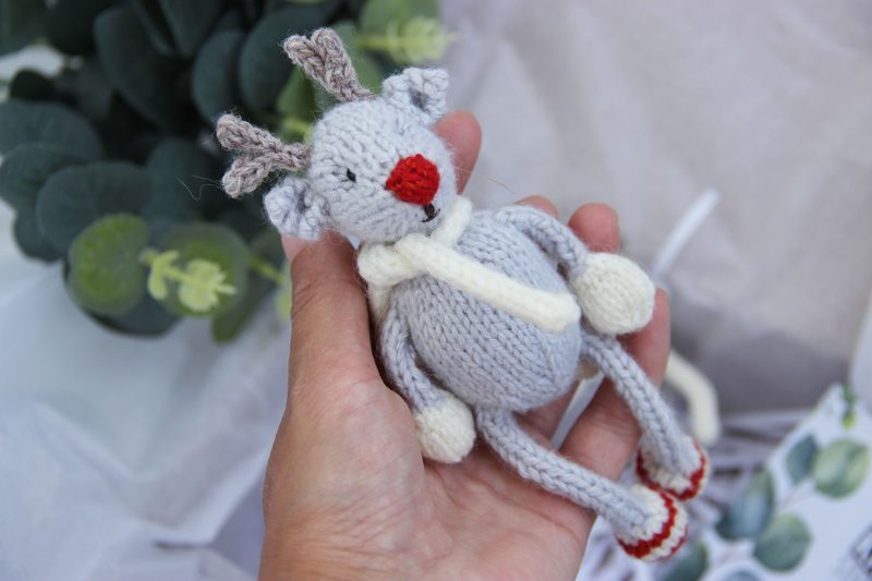 A small, knitted reindeer toy is being held in a hand, showcasing its light gray body and contrasting white scarf. The reindeer has a red nose and small brown antlers, with limbs and feet also knitted in gray. The background features soft, neutral-colored fabric and some green foliage, creating a cozy and festive atmosphere. The overall composition highlights the details of the toy while emphasizing its charming, handmade quality.