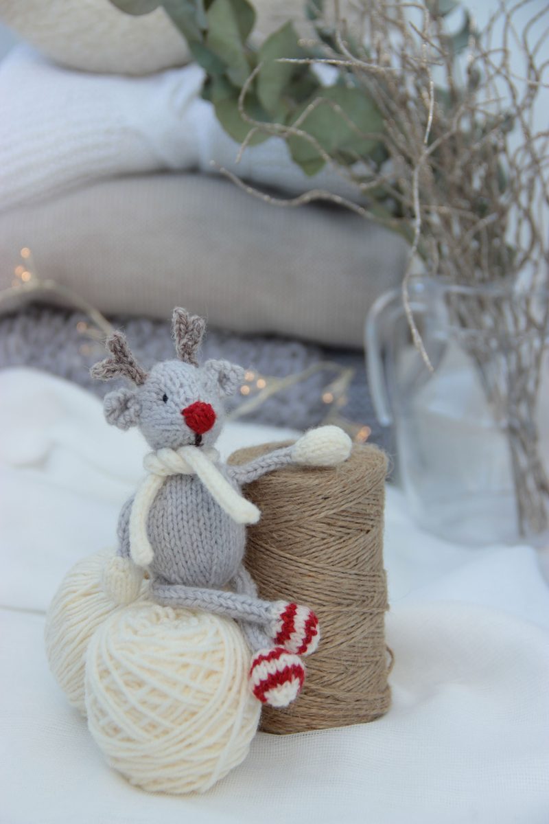 A knitted reindeer toy in grey and cream sits atop two yarn balls, one white and one tan, with red and white striped feet. The reindeer has small antlers and a red nose, wearing a cream scarf. In the background, there are cream blankets and soft greenery, with twinkling lights adding a festive touch. The image is softly lit, creating a cozy atmosphere.