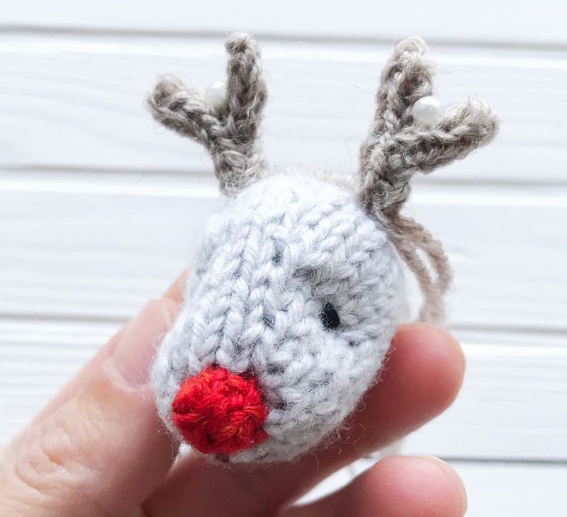 A close-up image of a small knitted reindeer toy, held in a person's hand. The reindeer features a gray body with a red nose and small black eyes. Its antlers are made from light brown yarn, decorated with two small white beads. The background consists of a white wooden surface, creating a clean and simple presentation.