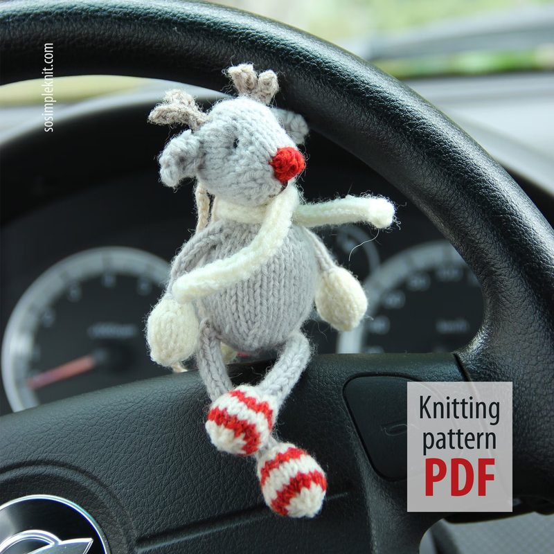 A knitted reindeer toy hangs from the steering wheel of a car. The reindeer is gray with a round body, white antlers, and a red nose. It has red and white striped legs and arms. The background shows a blurred dashboard with circular gauges, and a white square with the text "Knitting pattern PDF" in red. The overall scene is bright and focused on the reindeer, drawing attention to its playful design.