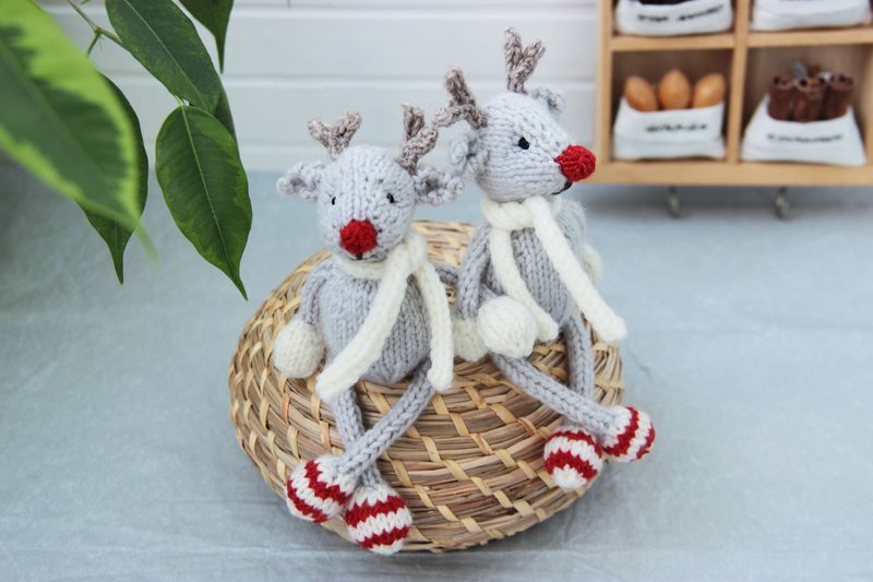 Two knitted reindeer toys, one slightly larger than the other, sit atop a round woven basket. The reindeer are mostly gray with red noses, and they have small, knotted antlers. Each is wearing a white scarf and has red and white striped legs. The background features a light-colored surface and a wooden shelf with small containers, adding a warm touch to the setting. A green plant is visible in the upper left corner, providing a contrast to the cozy scene.