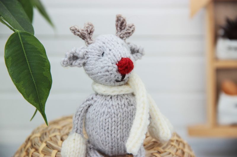 A small, knitted reindeer toy is seated on a woven basket. The reindeer features a light gray body, a red nose, and two antlers made of brown yarn. It is wearing a cream-colored scarf and has white mittens on its front paws. In the background, there is a green leafy plant to the left and a shelf with soft-colored items to the right, all set against a light, neutral backdrop.