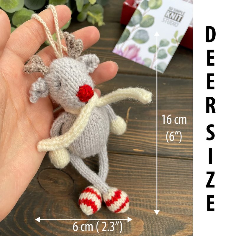 A hand holds a small, knitted reindeer toy with a gray body, red nose, and white antlers. The toy has striped red and white legs and a neutral-colored scarf. A ruler next to the toy indicates its size: 6 cm (2.3 inches) in width and 16 cm (6 inches) in height. The background features a blurred card with a floral design, set against a dark wooden surface, enhancing the focus on the reindeer.