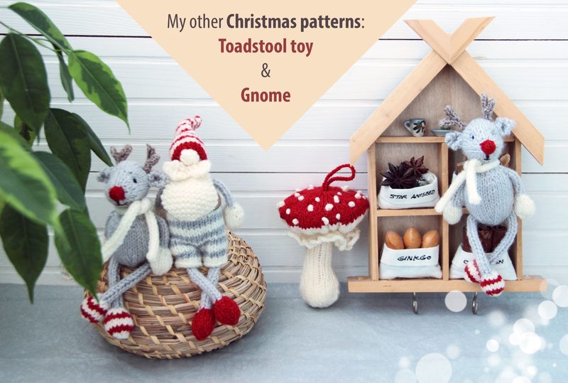 The image features two knitted reindeer toys, one wearing a red and white striped hat and the other in a striped sweater, sitting on a woven basket. Behind them is a small wooden house-shaped display with decorative elements, including a toadstool toy and small containers labeled with names. The background consists of white wooden panels, and there are green leaves from a potted plant on the left side, creating a cozy and festive atmosphere. The overall color scheme includes soft reds, whites, a