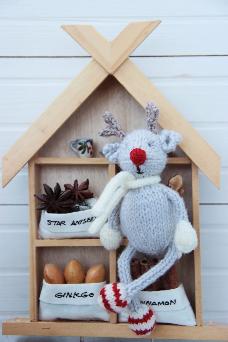 A knitted reindeer toy, featuring grey yarn for the body, white yarn for the paws, and a bright red nose, is posed in front of a small wooden display resembling a house. Behind the reindeer, there are shelves containing star anise, ginkgo nuts, and cinnamon, each labeled with small white fabric bags. The background consists of white wooden planks, and the roof of the display has a triangular design. The overall color scheme includes earthy tones with pops of red and white.