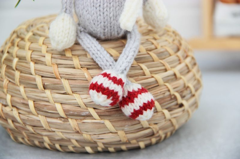 A close-up image of a knitted reindeer toy sitting on a textured, round wicker basket. The reindeer features a gray body with white ears and red-and-white striped legs, showcasing intricate knitting detail. The background is softly blurred, emphasizing the toy's colors and textures.