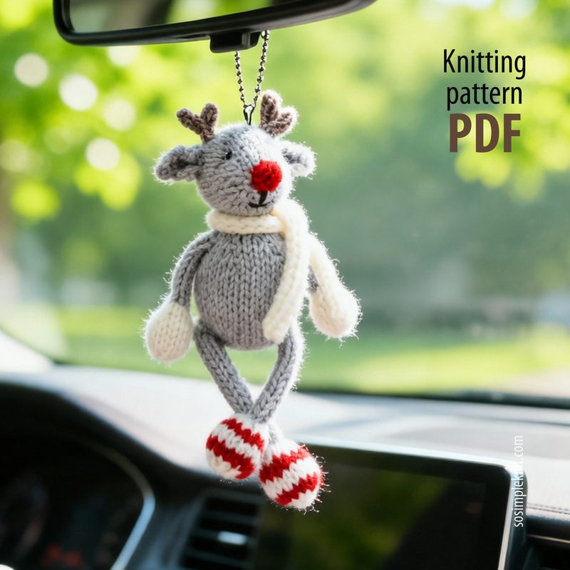 A small, knitted reindeer toy dangles from a rearview mirror, featuring gray yarn for the body, white yarn for the scarf, and red-and-white striped yarn for the feet. The reindeer has a red nose, large ears, and small antlers. The background shows a bright, blurred outdoor scene, suggesting daylight, while the focus remains on the reindeer. On the right side of the image, there is text labeled "Knitting pattern PDF."