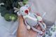 A small, knitted reindeer toy is being held in a hand, showcasing its light gray body and contrasting white scarf. The reindeer has a red nose and small brown antlers, with limbs and feet also knitted in gray. The background features soft, neutral-colored fabric and some green foliage, creating a cozy and festive atmosphere. The overall composition highlights the details of the toy while emphasizing its charming, handmade quality.