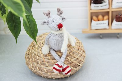 A knitted reindeer toy sits atop a woven basket. The reindeer has a light gray body with antlers and a red nose, wearing a white scarf. Its arms and legs are white, with red-and-white striped booties. In the background, there is a blurred green plant on the left and a wooden storage unit displaying small white bags filled with items, creating a cozy atmosphere. The setting features soft lighting, enhancing the warm, festive vibe of the scene.