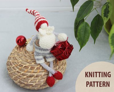 The image features a knitted gnome characterized by a red and white striped hat, sitting on a round woven basket. The gnome is holding a small red gift box and has a grey and white striped outfit. In the background, there are green leaves from a potted plant, and the bottom right corner includes text that reads "KNITTING PATTERN" in a light-colored circle. The overall color palette incorporates warm tones with a soft, cozy appearance.