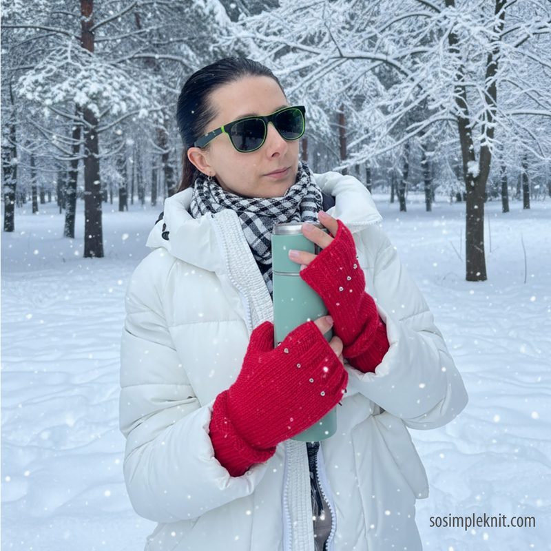 A woman wearing black sunglasses and a white puffy jacket stands in a snowy forest, holding a light green thermos. She has fingerless red gloves with a textured design and holds the thermos close to her chest. A black and white checkered scarf is visible around her neck. The background features snow-covered trees and a gentle snowfall, creating a winter atmosphere.